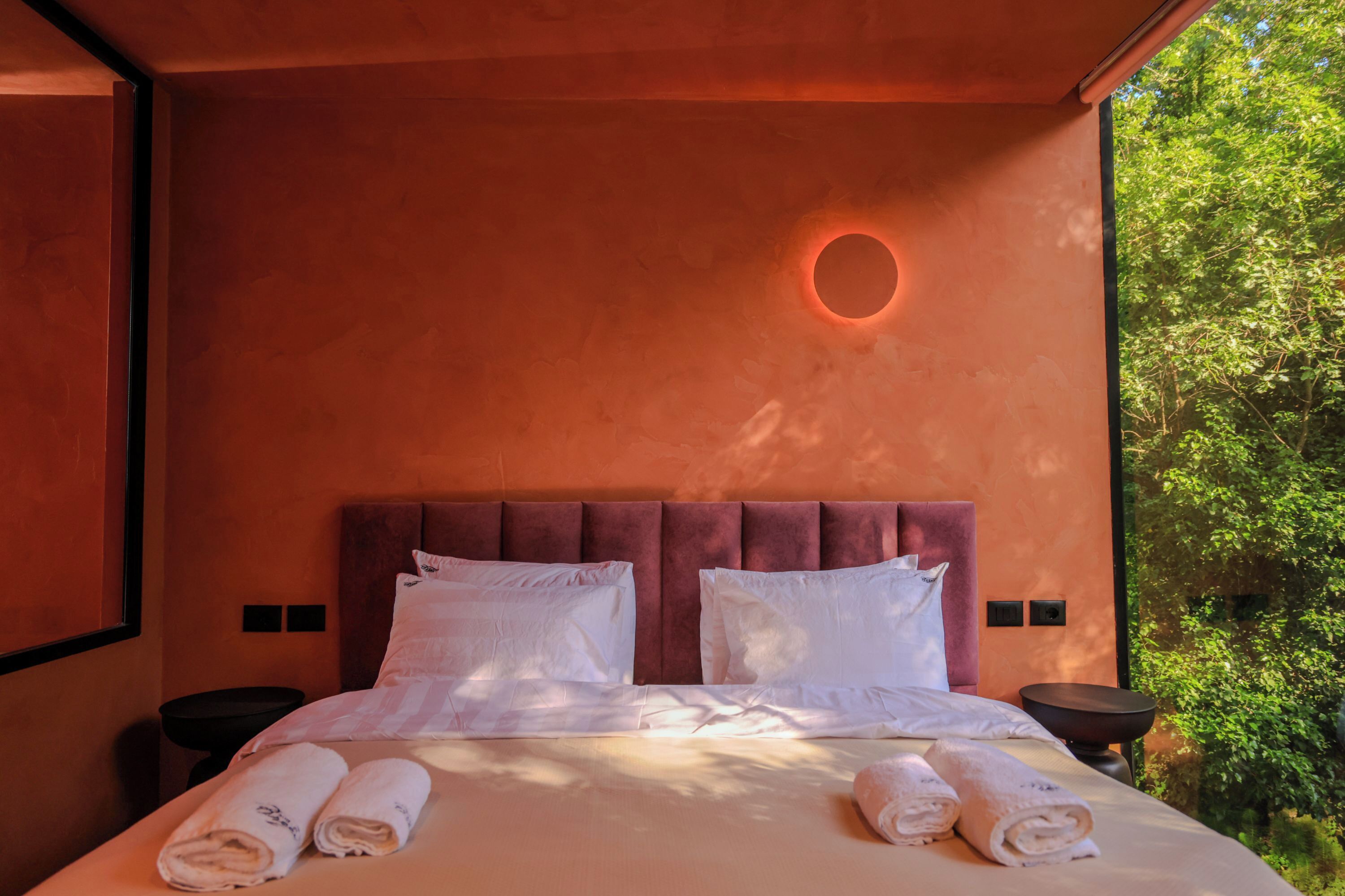 Une chambre avec un grand lit, deux oreillers, une applique murale ronde et une vue sur la verdure par une fenêtre.