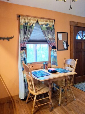 A wooden dining table with four chairs, a bulletin board with papers, and a window with patterned curtains.