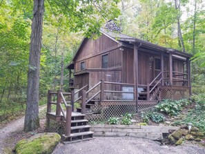 A wooden cabin with a wooden staircase leading up to a porch, surrounded by trees and a gravel path. - Old Man's Haven - Cabin 4 (Logan)