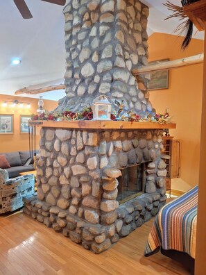 Una chimenea de piedra con repisa de madera, rodeada por una sala de estar acogedora con un sofá y un sillón estampado.