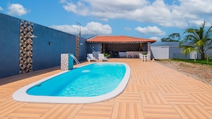 A swimming pool with a tiled deck, a covered seating area, and a blue sky. - Casa com Wifi Churrasqueira e Tima Localiza o (Camaçari)