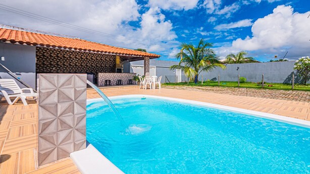 A swimming pool with a tiled deck, a covered seating area, and a fenced garden with palm trees.