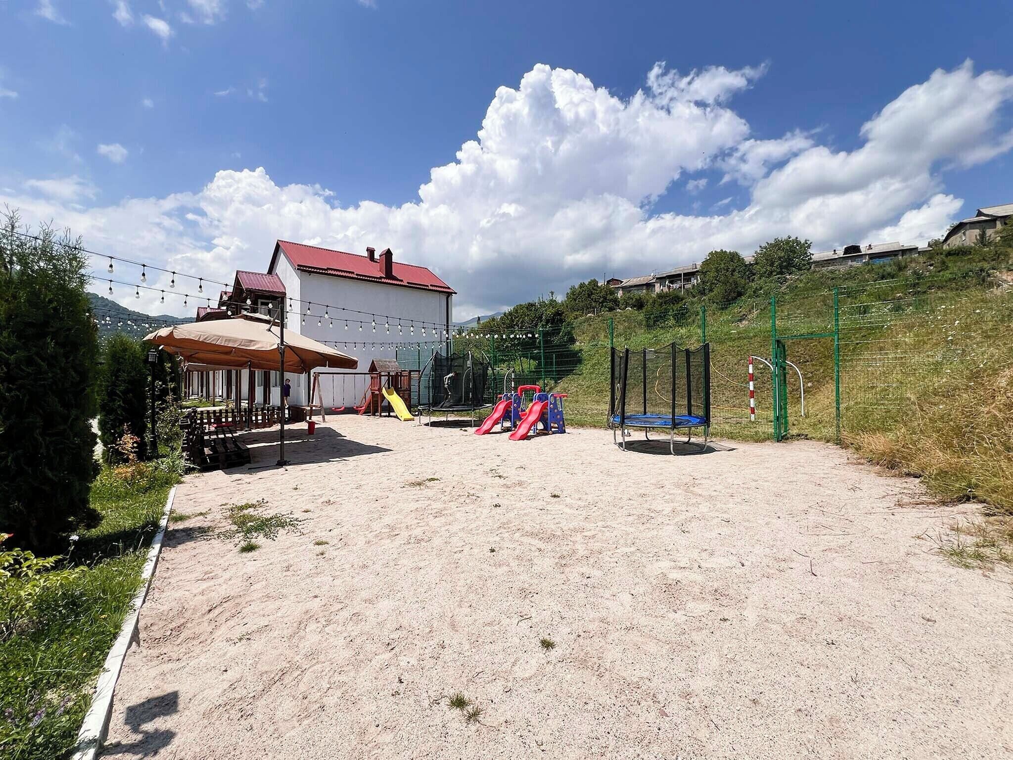 Zona de jocs amb tobogans i un trampolí, envoltada per una tanca i un edifici amb teulada vermella.