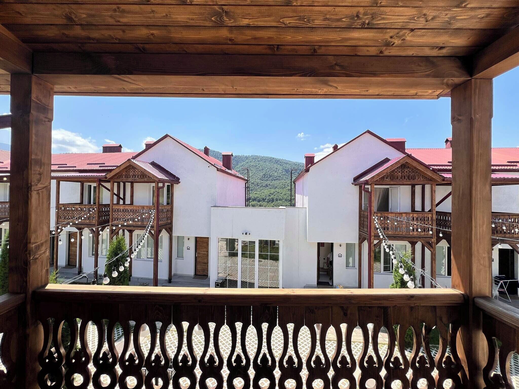 A wooden balcony with a view of two white buildings with red roofs, a wooden fence, and a mountainous background.