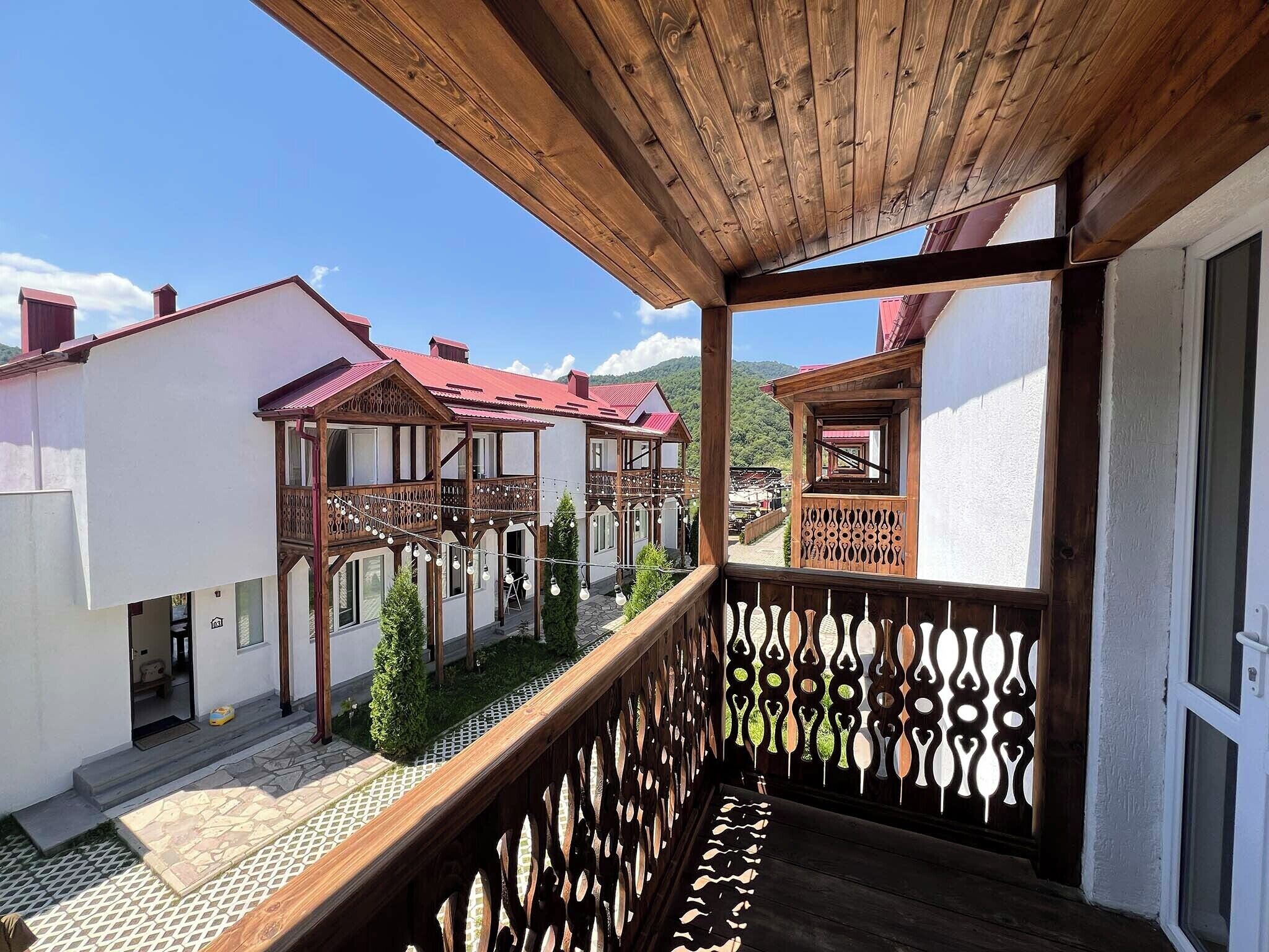 A wooden balcony with a patterned railing, overlooking a courtyard with traditional buildings and a clear sky.