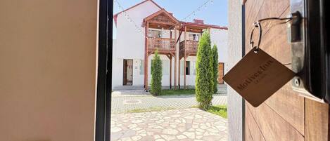 A view through a doorway to a courtyard with a stone-tiled floor, a wooden building with balconies, and a sign hanging on the door.