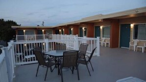 Un motel con un patio cubierto que incluye una mesa y sillas, barandal blanco y una hilera de habitaciones con ventanas.