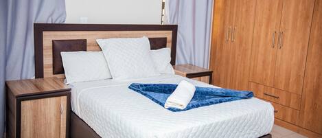 Una cama cuidadosamente hecha con colcha blanca y manta azul, cabecera de madera y armario de madera con tiradores plateados.