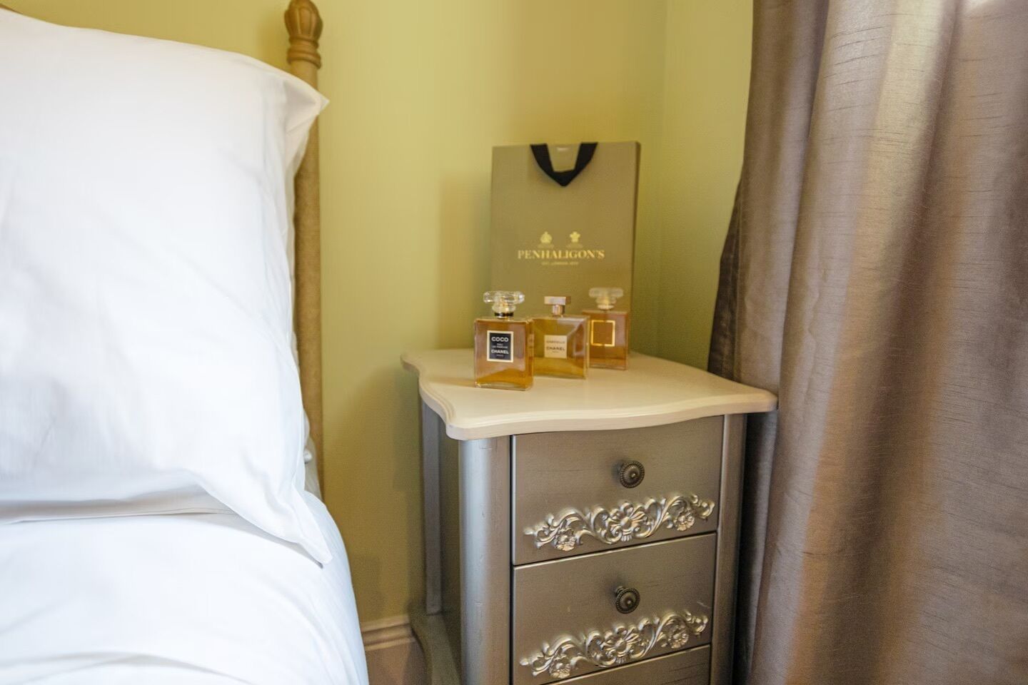 Un lit bien fait, avec du linge de lit blanc, une table de chevet sur laquelle se trouve un porte-stylos, et trois flacons de parfum posés dessus.