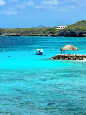 Une mer turquoise aux eaux limpides, avec un bateau et une construction à toit de chaume sur une petite île.