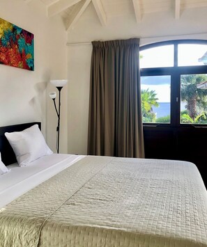 Habitación de hotel con cama, un cuadro en la pared, una lámpara de pie y vista al mar por la ventana.