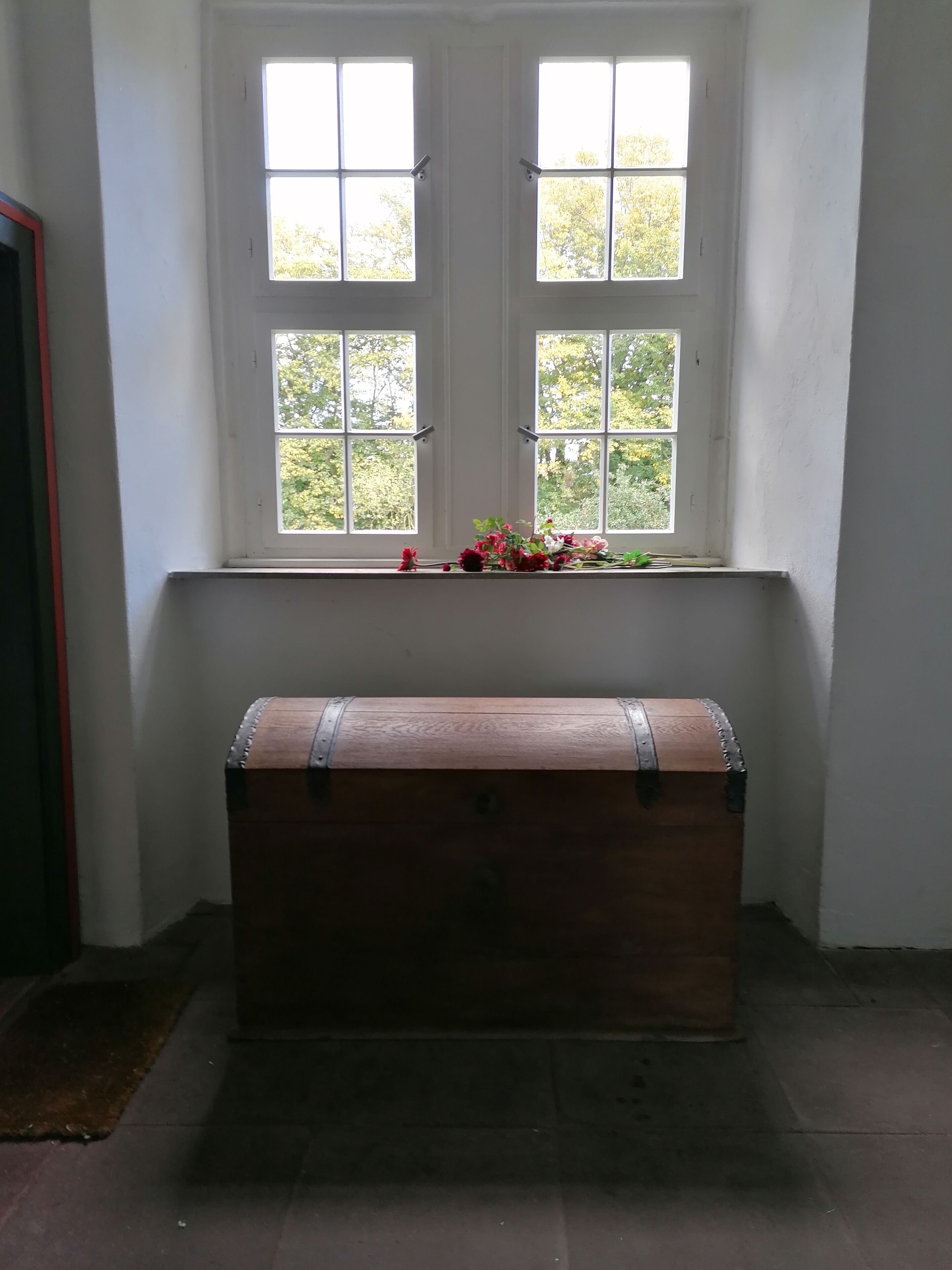 Ein Zimmer mit einem großen Fenster, einer hölzernen Truhe und einem kleinen Tisch mit Blumen.