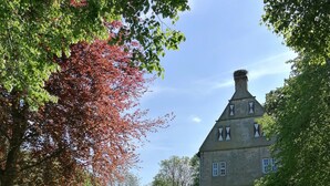 A historic stone building with a steeply pitched roof and multiple windows, surrounded by well-maintained hedges and trees with autumn foliage. - Apartment in medieval castle from the 14th century on the Weser cycle path (Petershagen)