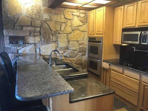 A kitchen with granite countertops, a stone backsplash, stainless steel appliances, wooden cabinets, and a built-in microwave.