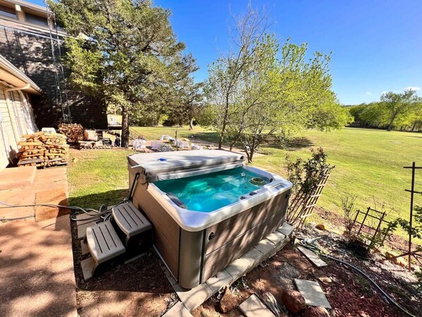 A hot tub in a backyard with a wooden deck, a pile of firewood, and a lawn.