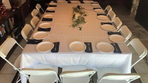 Une longue table à manger, dressée pour un repas, entourée de chaises blanches, avec un piano en arrière-plan et une cheminée.