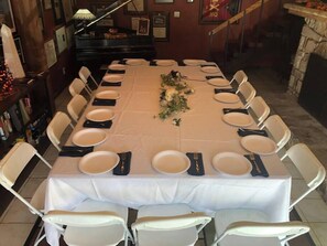 A long dining table set for a meal with white chairs, a piano in the background, and a fireplace.