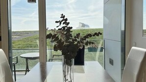 A modern dining area with a glass table and chairs, a vase with dried flowers, and a view of a distant landscape through large windows. - Bridge River Apartment @ Parque das Nações (Lisboa)