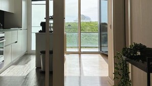 A modern kitchen with white cabinets, a dining area with a table, and a large sliding glass door leading to a balcony with a view of greenery. - Bridge River Apartment @ Parque das Nações (Lisboa)