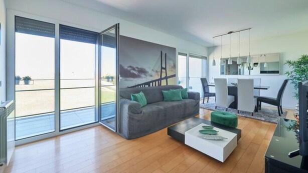 A modern living room with a gray sofa, a white coffee table, and a green ottoman. There is a large window with a view of a balcony and a painting on the wall. - Bridge River Apartment @ Parque das Nações (Lisboa)
