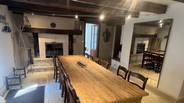 Une longue table à manger en bois, entourée de chaises, une cheminée en pierre et un miroir qui reflète la pièce.
