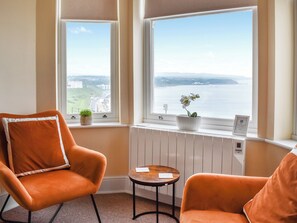 A room with two orange chairs, a small table, and a view of a body of water through large windows.