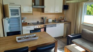 Une cuisine compacte équipée d’un réfrigérateur, d’une cuisinière et d’un lave-vaisselle. On y trouve une table à manger avec des chaises et un canapé. Une fenêtre apporte de la lumière naturelle.