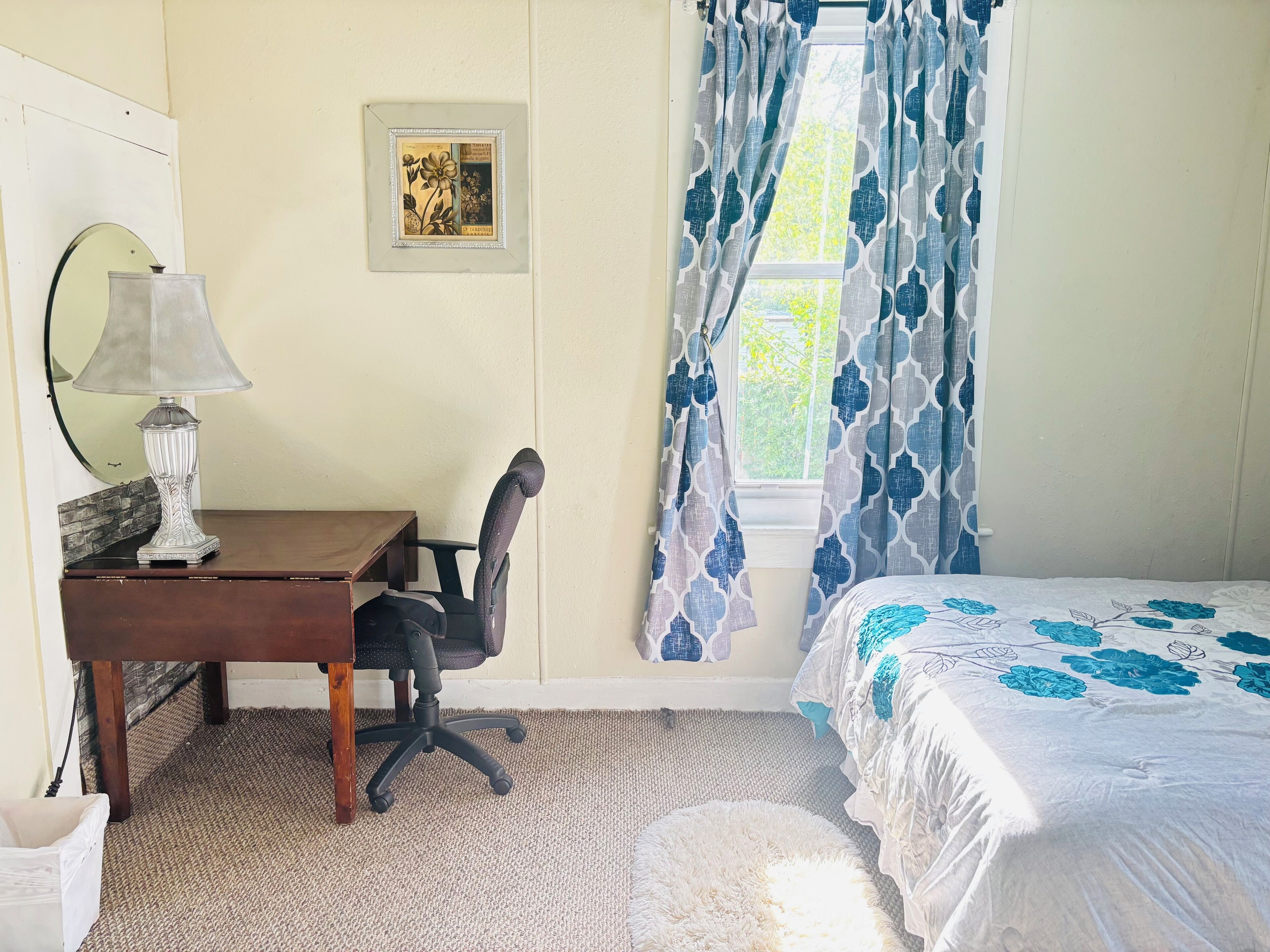 Een slaapkamer met een bed, bureau met lamp, stoel, een inlijsting afbeelding aan de muur, en een raam met gordijnen.