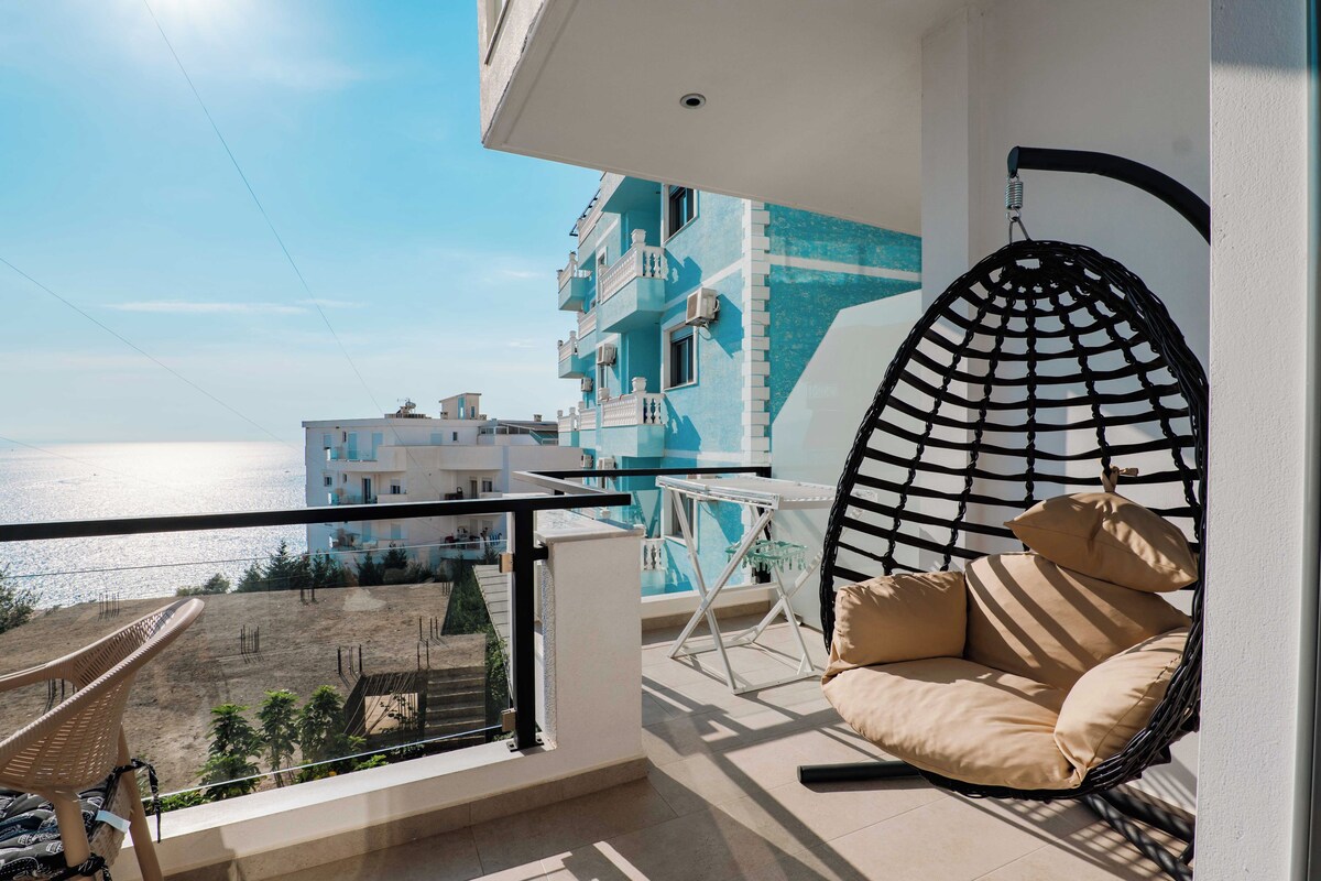 A balcony with a hanging chair, a small table, and a view of the sea and buildings.