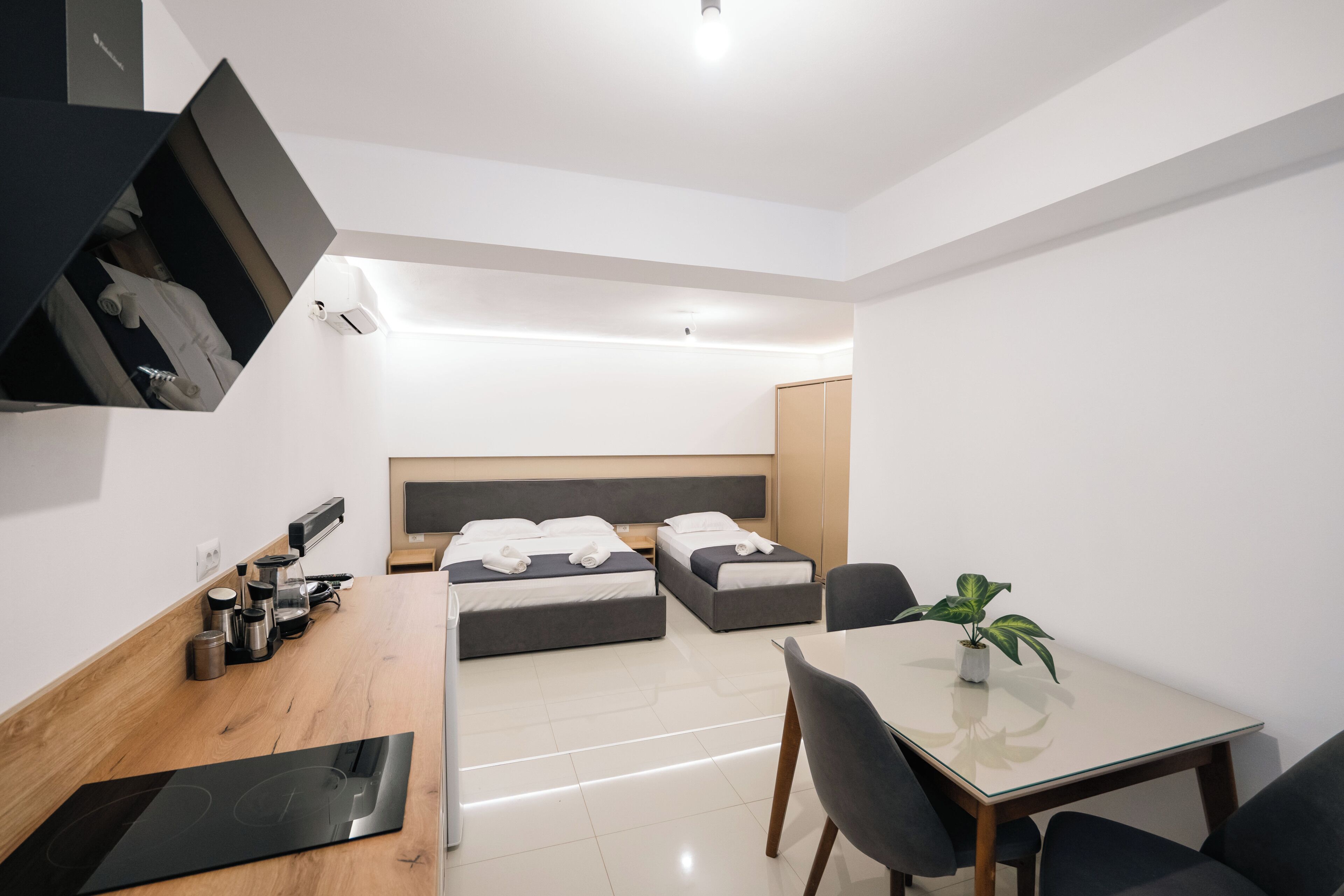 A modern hotel room with two beds, a dining table, a kitchen area with a stove, and a small plant on the table.