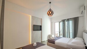 Camera d'albergo con due letti, una TV a schermo piatto fissata al muro, una lampada a sospensione moderna e una finestra con tende.