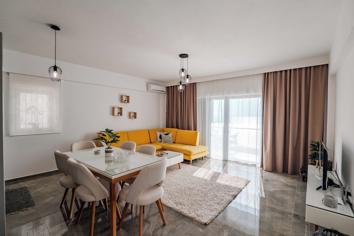 Una sala de estar moderna con una mesa de comedor de vidrio, sillas color beige, un sofá amarillo y un ventanal con cortinas.