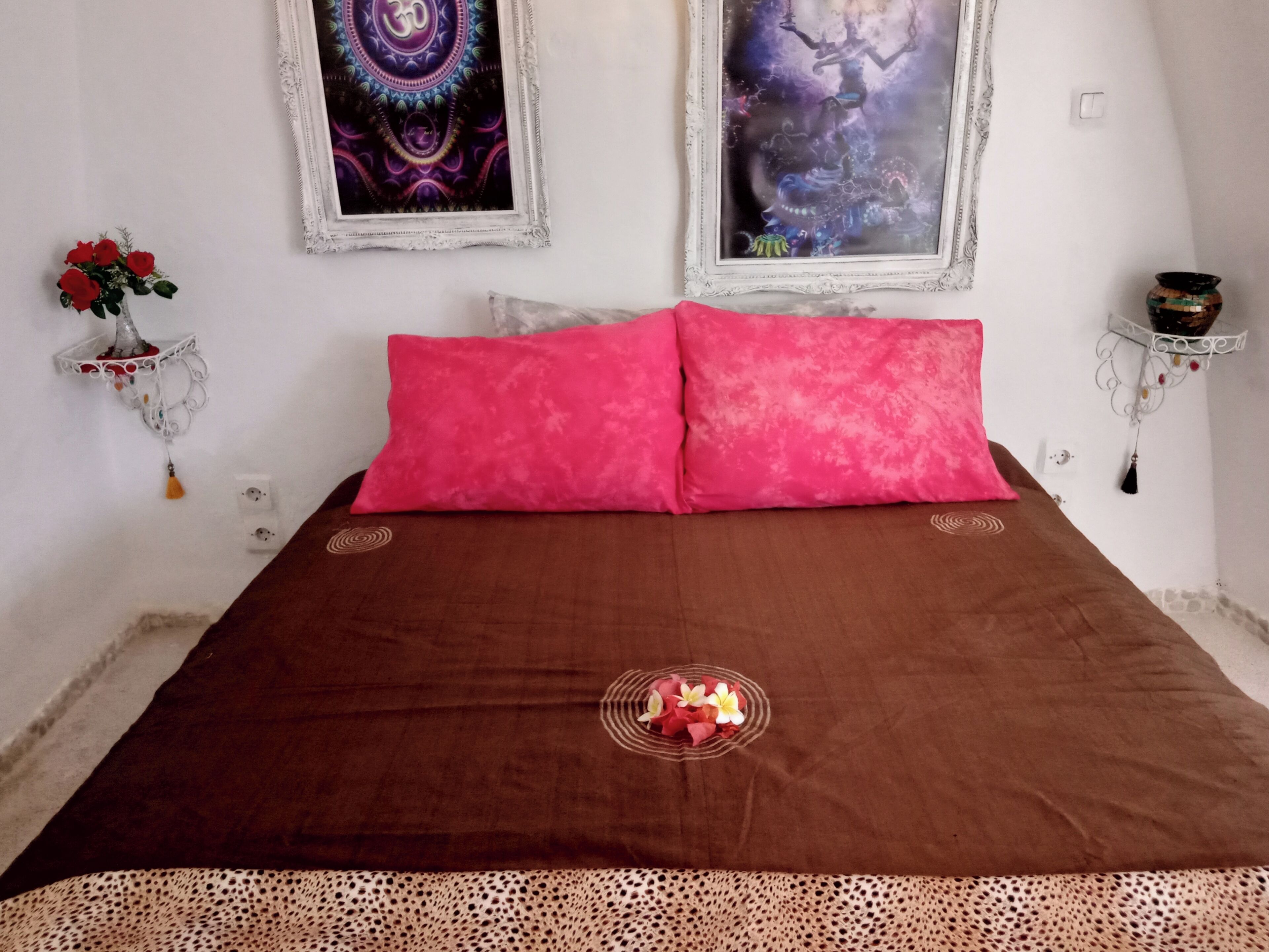 Un dormitorio con una colcha marrón, dos almohadas rosas, un cojín decorativo con flores y dos cuadros colgados en la pared.
