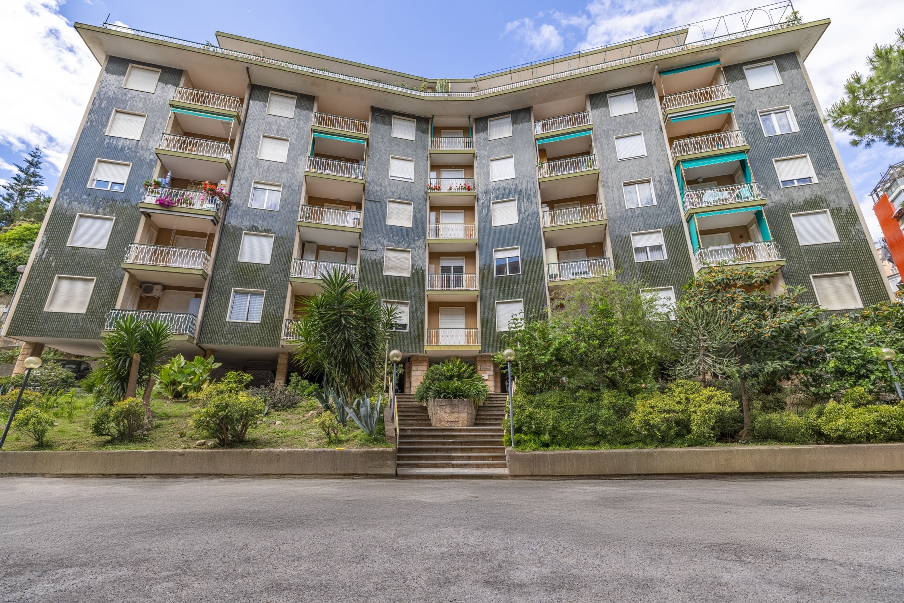 A multi-story residential building with balconies, a central entrance, and a landscaped garden area.