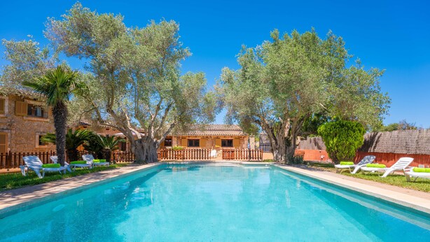 Une piscine entourée de chaises longues, bordée d’arbres et située à proximité d’un bâtiment dont le toit est recouvert de tuiles.