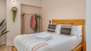 A neatly made bed with a wooden headboard, two pillows, and a folded white towel. A small plant hangs from the wall, and a wooden rack with a pink garment and a green towel is visible. - Marconia Boutique Residence   Dúplex Premium (Girona)
