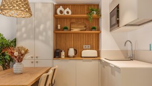 A compact kitchen with wooden shelves, a built-in oven, a sink, and a dining area with a table and chairs. - Marconia Boutique Residence   Dúplex Premium (Girona)