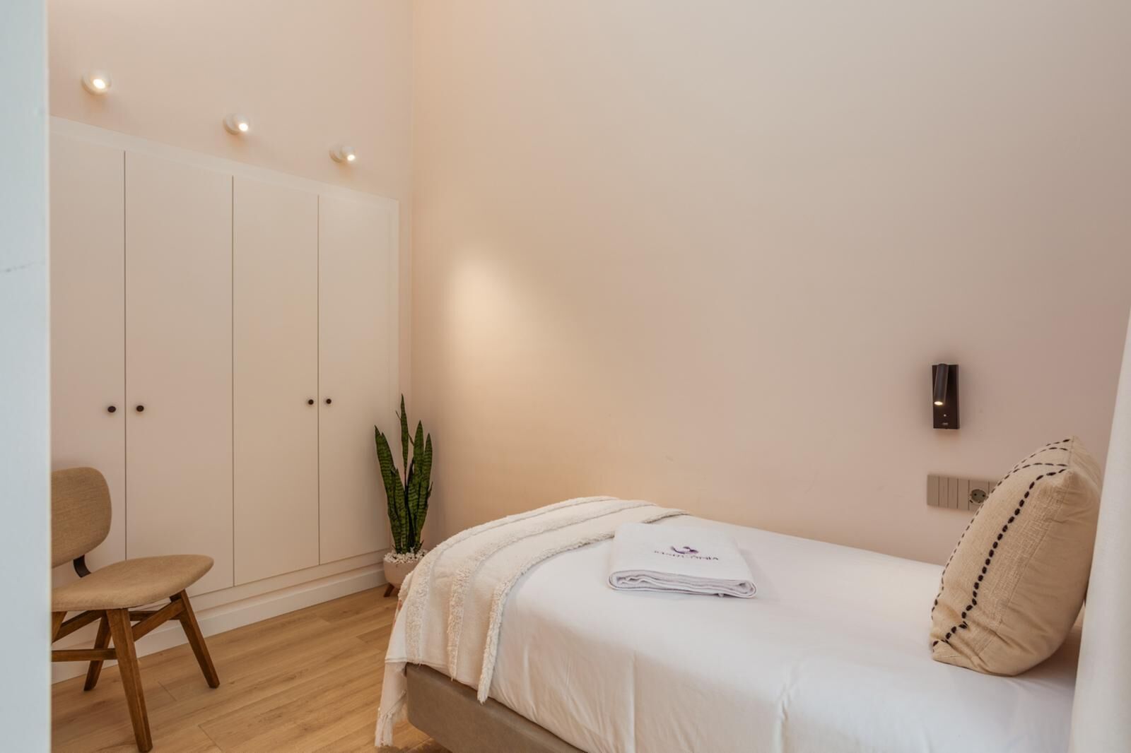A bedroom with a bed, a chair, a wardrobe, a plant, and a wall-mounted light fixture.