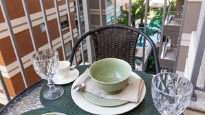 Una mesa de comedor con un posavasos verde, un bol verde, un vaso transparente y una cuchara plateada en un balcón con vistas a un edificio y palmeras.