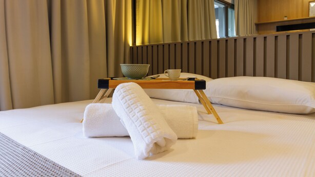 Una cama bien tendida con sábanas blancas, una mesita de noche de madera con un cuenco y una taza, y una ventana con cortinas.