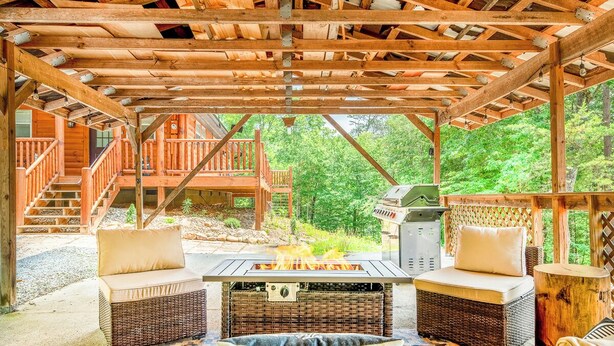 Un espacio al aire libre cubierto con zona de fogata, muebles de mimbre y vistas a una terraza de madera y bosque.