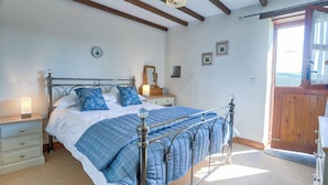 A bedroom with a metal bed frame, a bedside table, a lamp, and a view of the outside through a wooden door. - Clover Cottage (Saint Wenn)