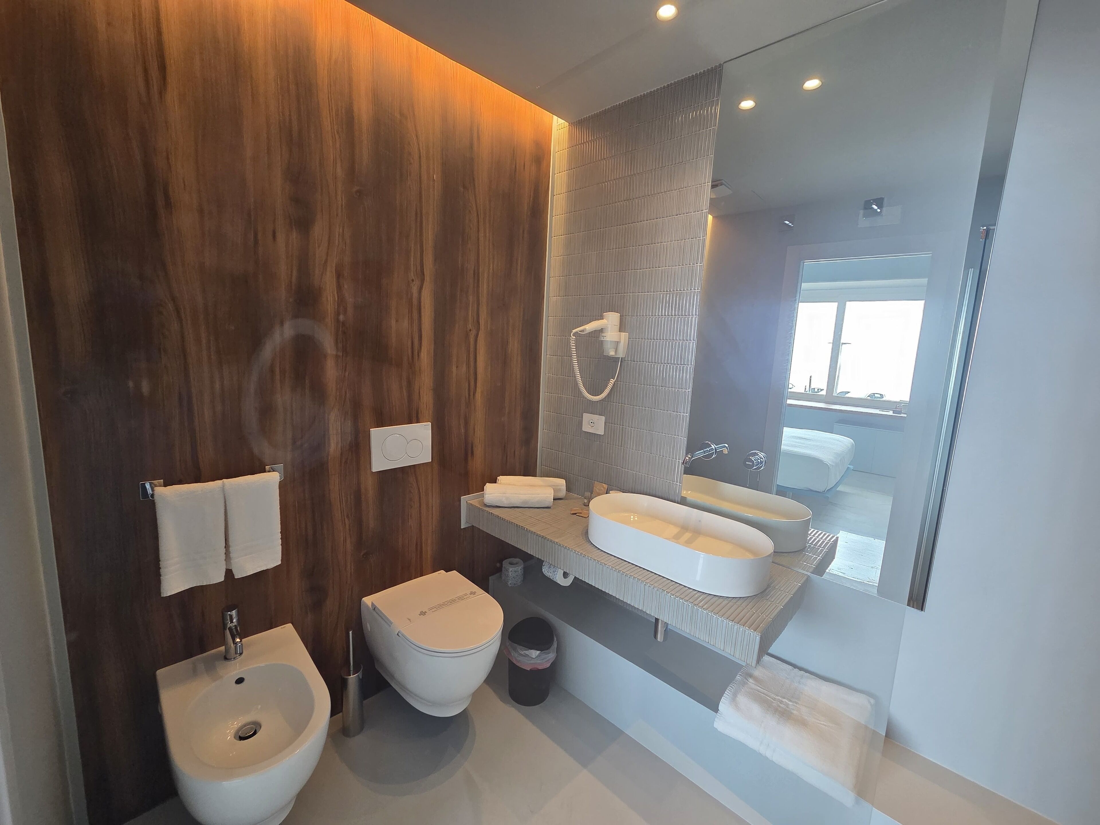 A modern bathroom with a wooden wall, a large mirror, a bidet, a toilet, and a sink.