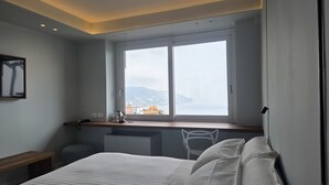 A hotel room with a bed, a desk with a chair, a window with a view of a cityscape, and a mounted picture on the wall. - TPH Rooms (Taormina)