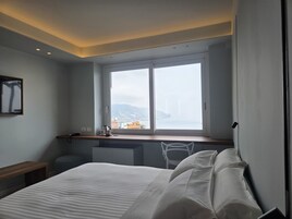 Quarto de hotel com uma cama, uma escrivaninha com cadeira, uma janela com vista para a cidade e um quadro pendurado na parede.