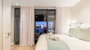 A hotel room with a large bed, a view through a sliding door, and a balcony with a table and chairs.