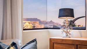 A room with a view of mountains through a window, a lamp on a wooden nightstand, and a sofa with cushions.