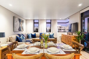 Una sala de estar moderna con comedor, un conjunto de sofá, una mesa de comedor de madera y un ventanal con vistas a edificios.