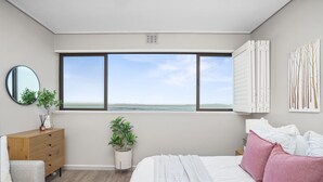 A bedroom with a large window, a bed, a dresser, a mirror, and a plant. - Searenity Tides - Two Bedroom Apartment, Sleeps 4 (Cape Town)