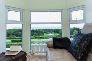 A room with a large bay window overlooking a garden and a distant view of a body of water.